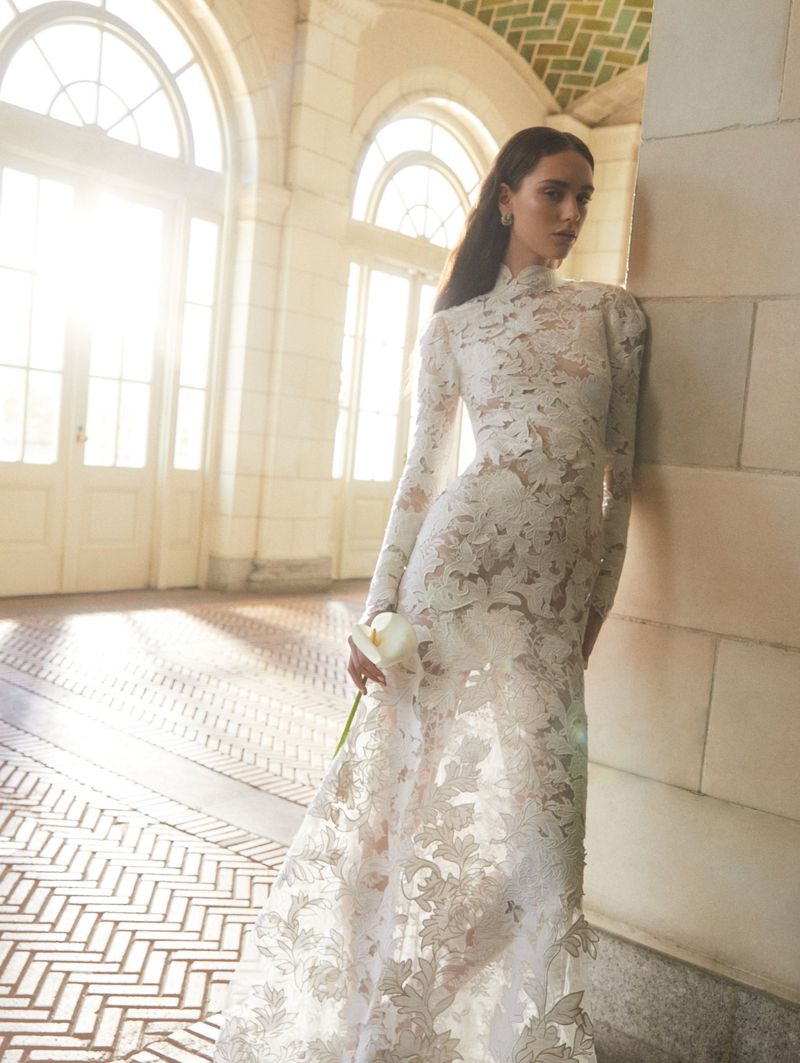 Elegant woman in a floral-lace gown holds a white flower in an indoor setting with large arched windows and herringbone floors.