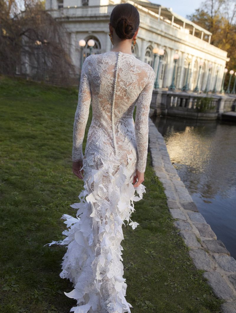 Back view of a woman wearing a white lace gown with a train in a park, walking towards a white building near a pond on a sunny day.