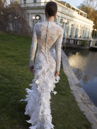 Back view of a woman wearing a white lace gown with a train in a park, walking towards a white building near a pond on a sunny day.