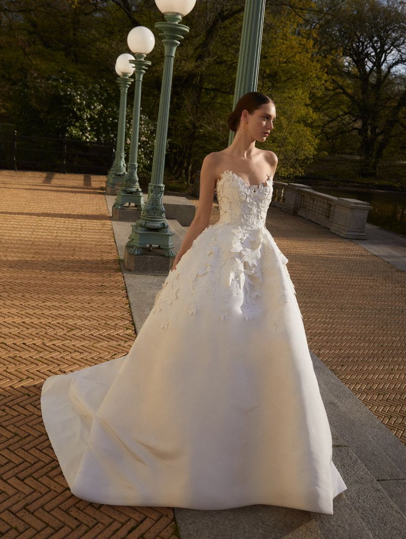 Woman in elegant, strapless white wedding gown with floral appliques poses outdoors on brick path with green trees and ornamental lamps.