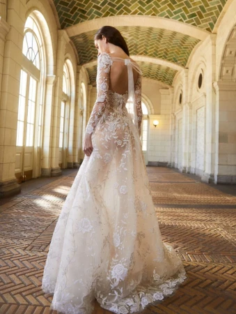 Woman in an ornate, lace-covered white wedding dress stands in a hallway featuring arches, windows, and intricate geometric tile work.