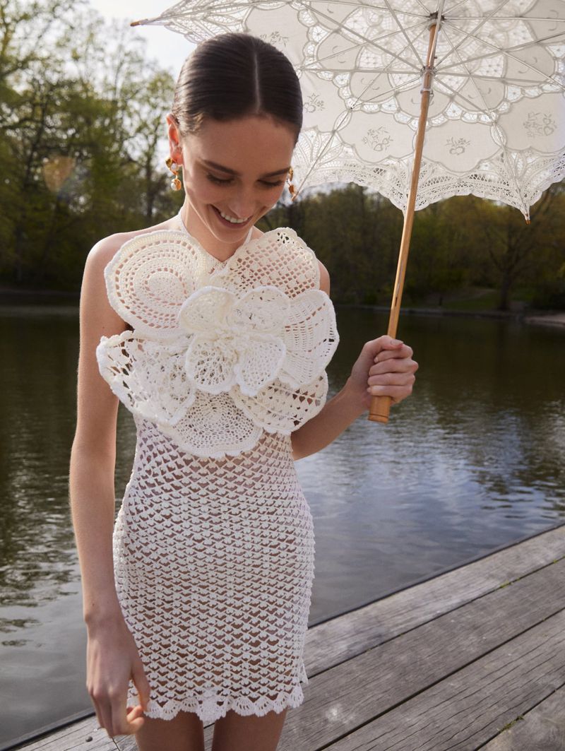 Woman in white crochet dress and holding an umbrella standing on a wooden deck by a lake surrounded by trees with green leaves.