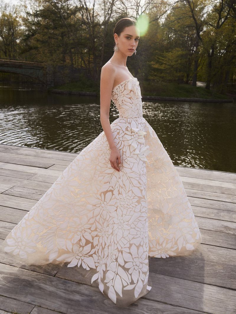 A woman stands on a wooden platform in a flowing floral patterned strapless gown with a waterfront and trees in the background.