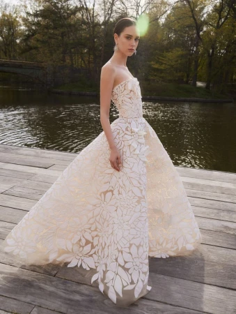 A woman stands on a wooden platform in a flowing floral patterned strapless gown with a waterfront and trees in the background.