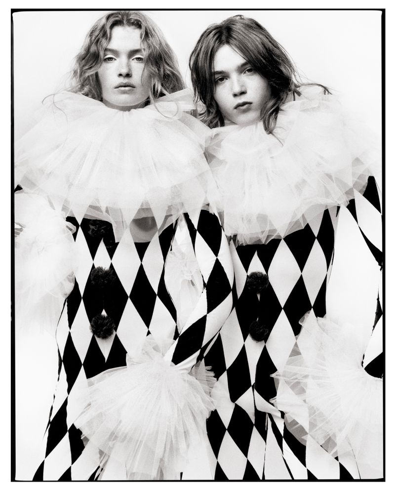 Striking black and white portrait of two people in matching diamond pattern outfits and white ruffled collars against a white backdrop.