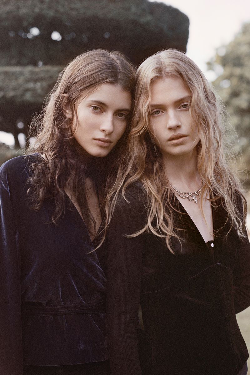 Two female models with long hair wearing dark tops outdoors against a green garden background