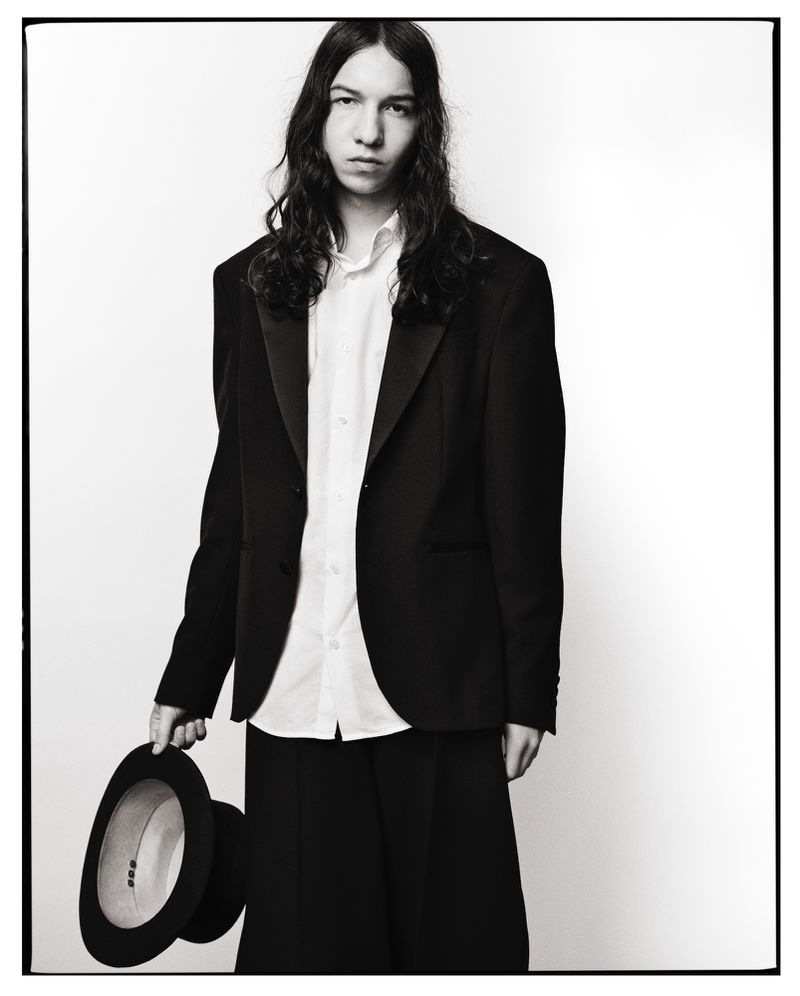 Portrait of a young adult in a black tuxedo jacket and white shirt, holding a black hat against a white background.