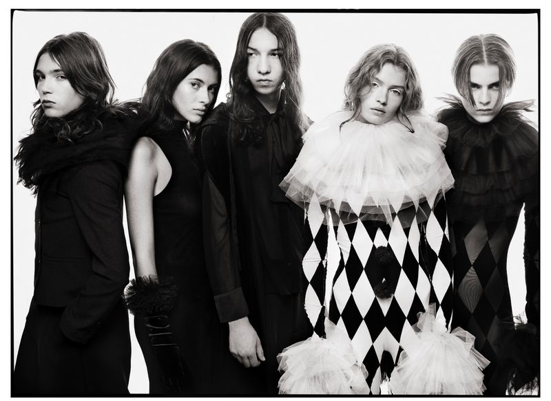 A black and white fashion photograph of five young adults in formal attire with ruffled necklines posing closely together in a studio.