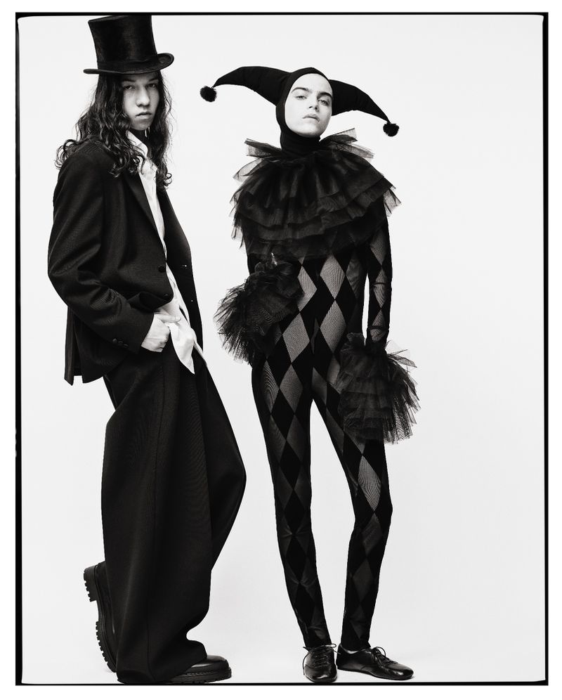 Two figures in stylized attire pose against a stark white backdrop, one in a black suit and top hat, the other in a harlequin costume.