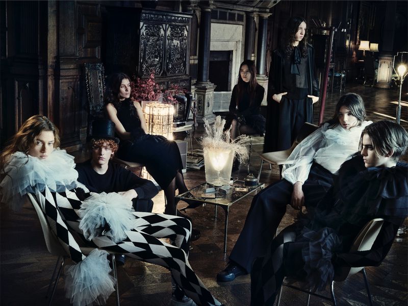 Group of seven people in a dark room with ornate walls, sitting in chairs. Some are wearing black and white harlequin outfits.