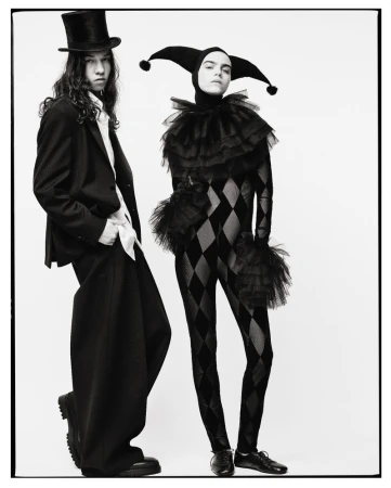 Two models wearing harlequin-inspired fashion against a plain white background, one in a top hat and suit, the other in a jester hood and bodysuit