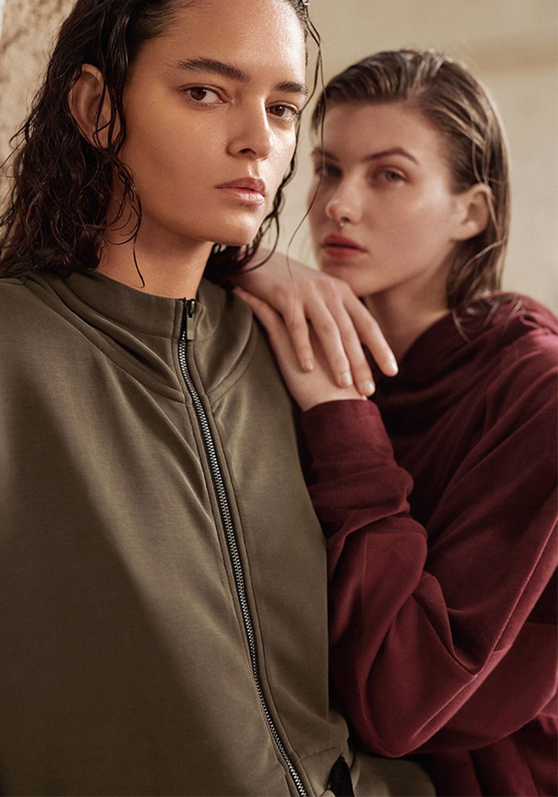 Two women pose in studio, wearing green zip-up and red hoodie. Wet hair, neutral expressions, indoor setting. Fashion, beauty, portrait photography.
