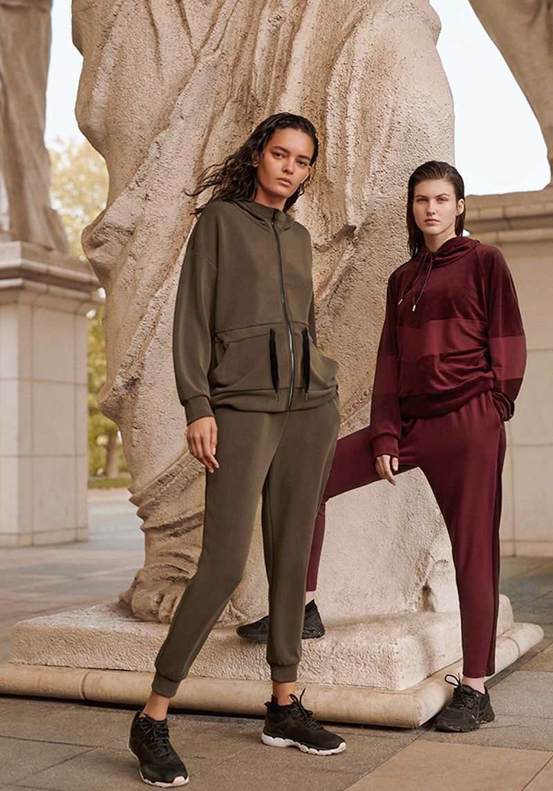 Two women pose near a sandstone sculpture wearing matching athleisure outfits consisting of hooded sweatshirts and track pants in a studio-like setting.