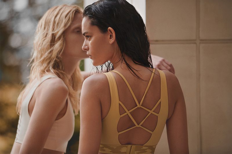 Two women in athletic tops. One woman with dark hair faces forward with the back of her yellow top visible, as another whispers nearby.