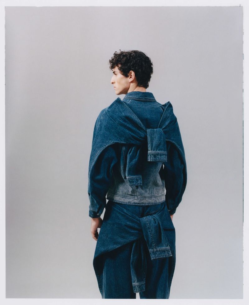 Back view of a person in a denim jacket and pants with a large bow detail on the back of the jacket in a studio setting.