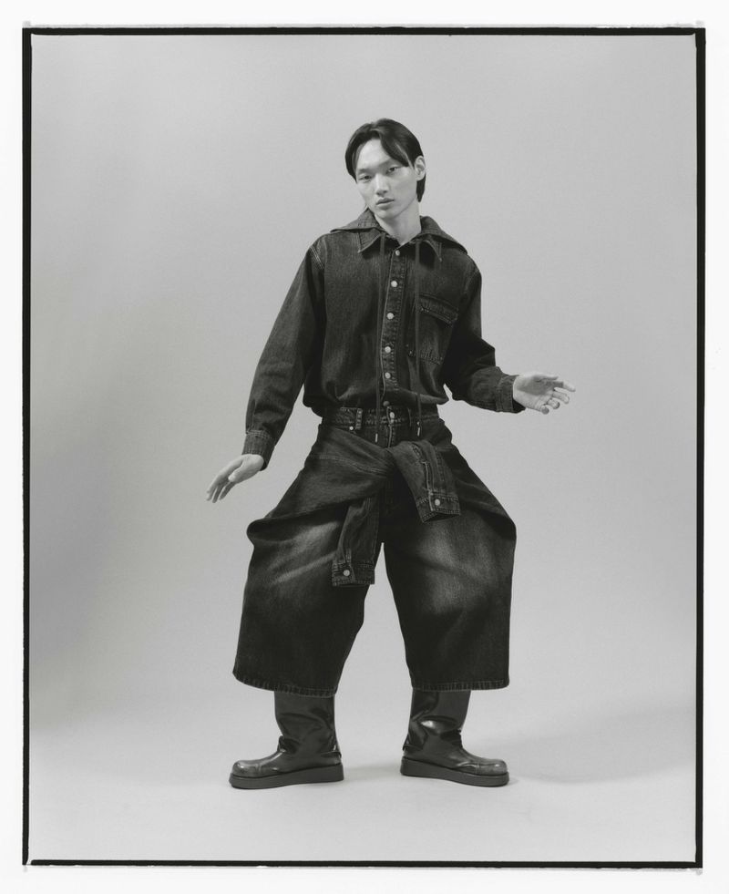 An Asian male model poses in a photo studio wearing an all-denim outfit including an oversized shirt, pants, and leather boots.