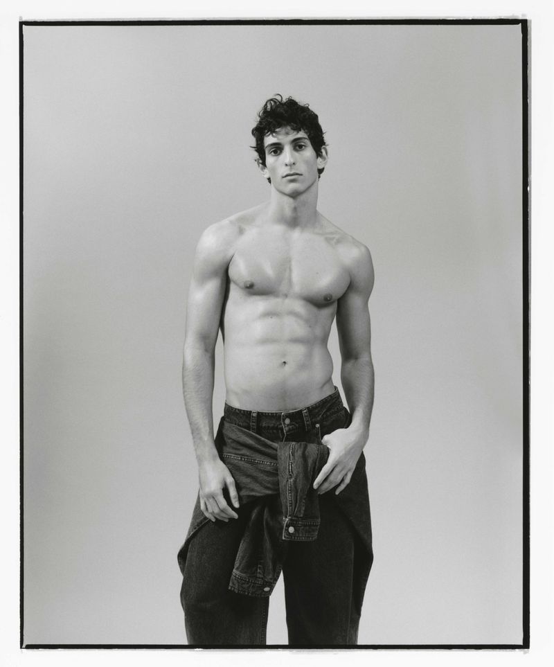 Monochrome portrait of a male fashion model with a muscular physique, posing shirtless in jeans with jacket tied around the waist in a studio.