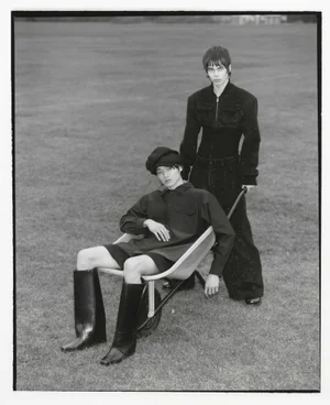Two models in dark avant-garde tailoring and tall leather boots pose with a wheelbarrow on an open grassy field in black and white.