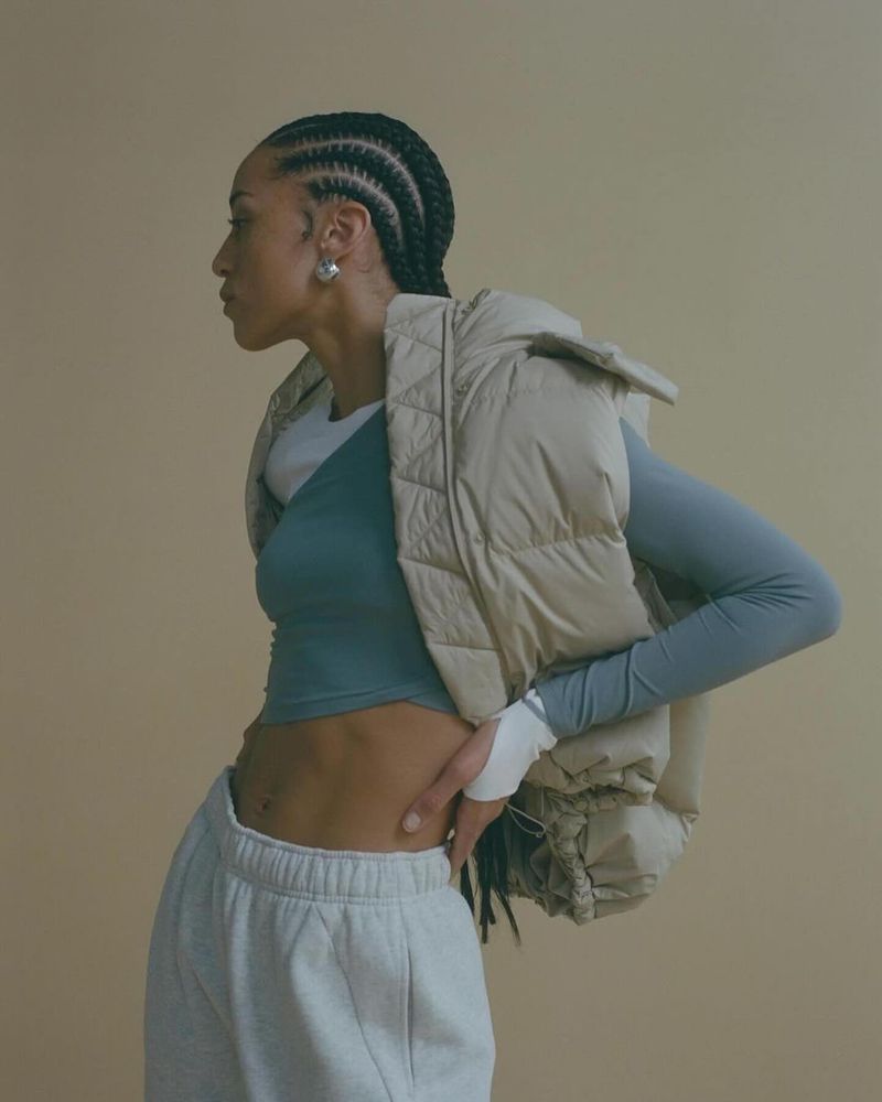 Athletic woman poses in sports bra, puffer vest and grey sweatpants in a studio with a neutral background.