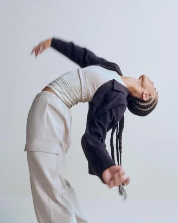 Dancer Alanna Archibald arching backward in cream cargo pants and a draped black jacket against a plain white studio backdrop.