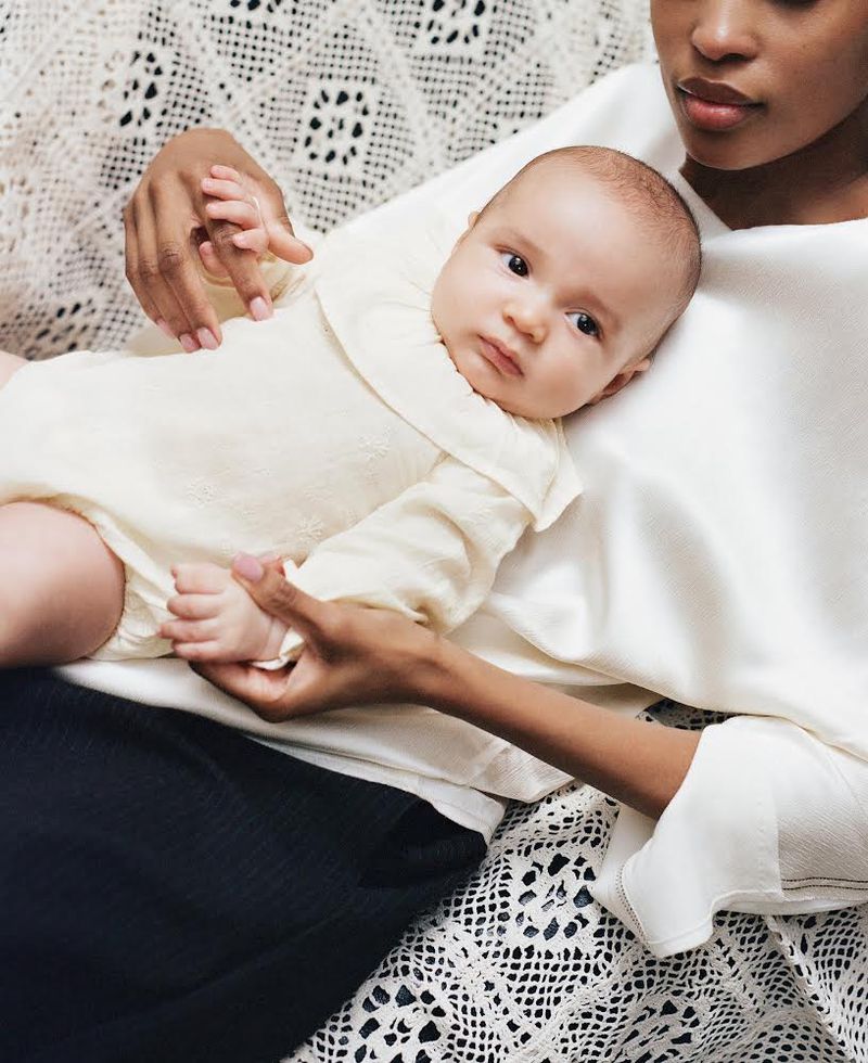 A mother gently cradles her infant, both adorned in light-colored clothing against a cream-colored, patterned surface, showcasing a tender moment.