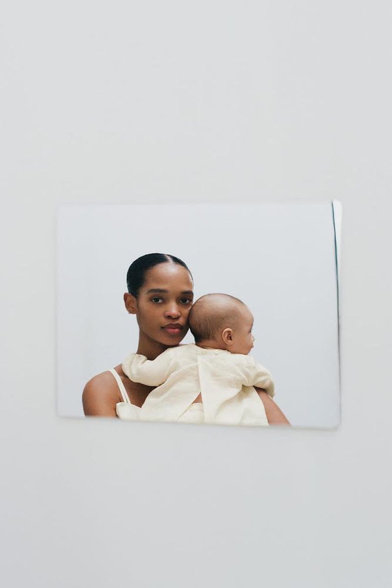 Woman with dark hair holding a baby in her arms, both wearing light colored clothing in a softly lit indoor setting.