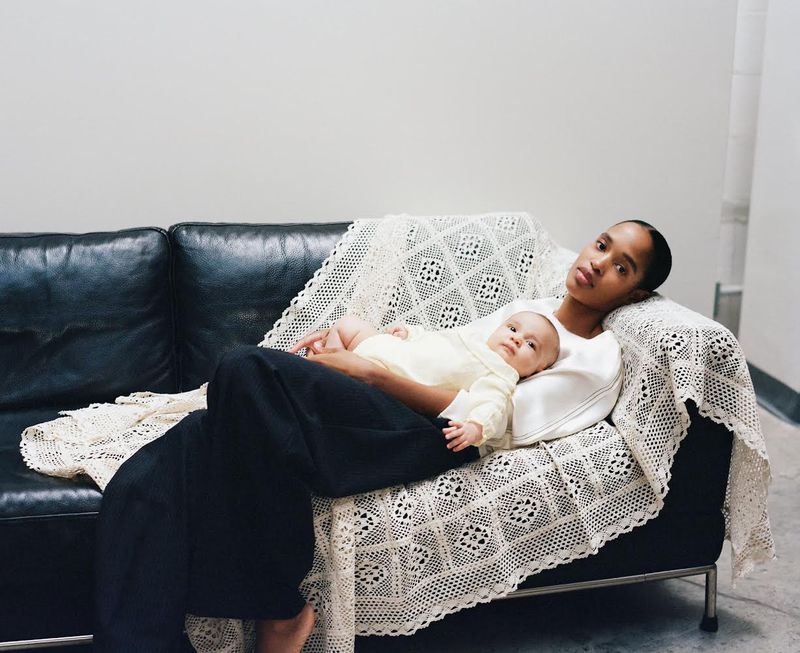 Young Black woman rests on a black leather sofa with her baby, draped in a lacy white blanket indoors against a grey wall.