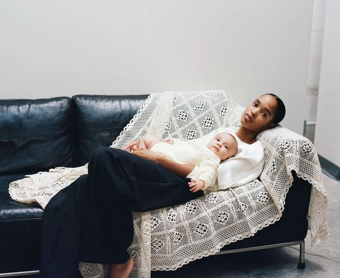 Model Marihenny Pasible reclines on a black leather couch draped with a white crochet blanket, holding a baby in a yellow knit outfit.