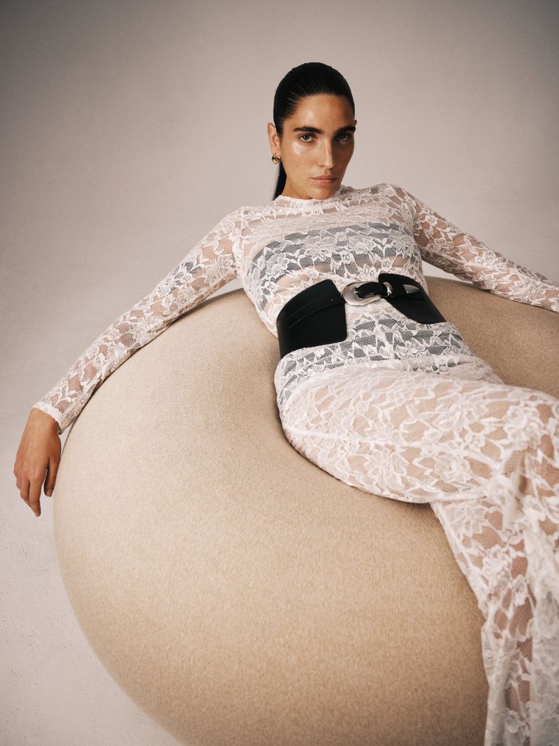 Adult woman with dark hair poses in a long-sleeved lace dress, black belt, and gold earrings while sitting on a circular fuzzy chair.