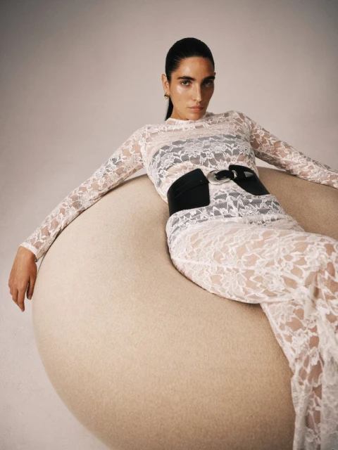 Adult woman with dark hair poses in a long-sleeved lace dress, black belt, and gold earrings while sitting on a circular fuzzy chair.