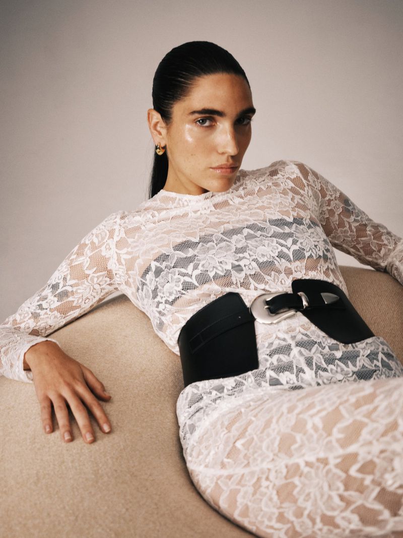 Fashion photo of a slender adult female reclining on a beige couch. She is wearing a white lace top, a matching skirt, and a wide black belt.