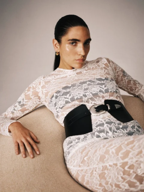 Fashion photo of a slender adult female reclining on a beige couch. She is wearing a white lace top, a matching skirt, and a wide black belt.