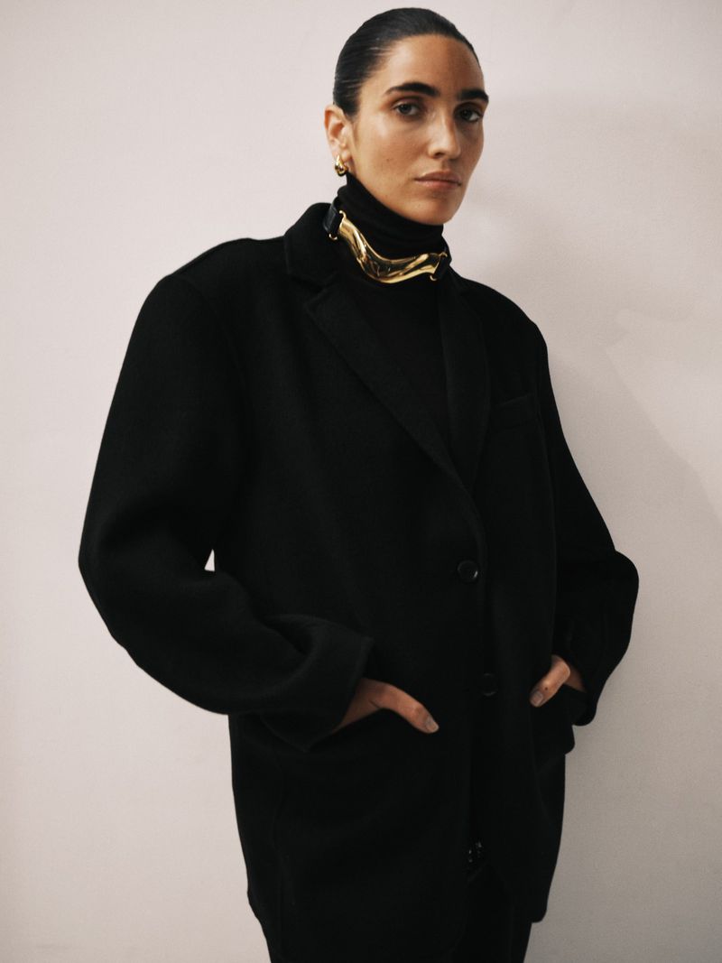Elegant portrait of a brunette woman with her hands in the pockets of a black wool coat over a black turtleneck top.