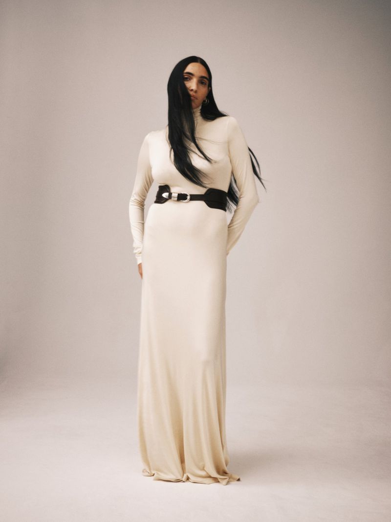 A full-length portrait of an adult female with long, dark hair, wearing a white dress and a black belt against a neutral backdrop.