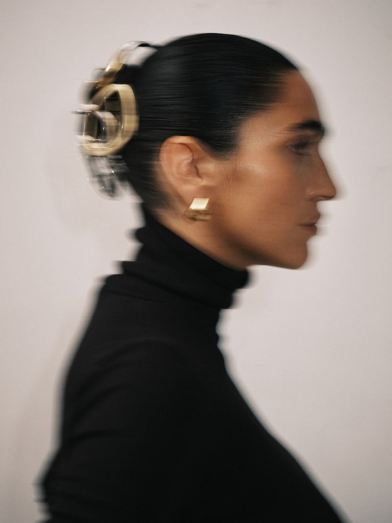 Woman in profile wearing a black turtleneck, gold earrings and a gold hair clip, with a blurred effect against a light grey background.