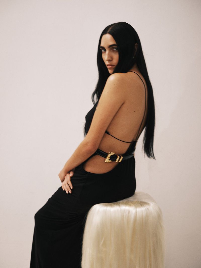 Brunette woman in backless black dress with belt seated on stool in a light studio setting looking at camera. Fashion photography.