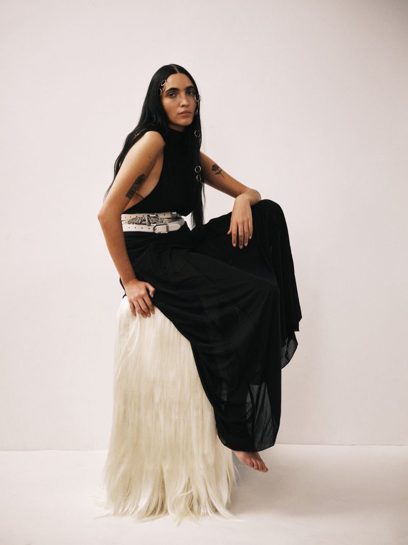 A stylish woman with long dark hair poses indoors, seated on a white fur stool, wearing a black dress with a wide white belt.