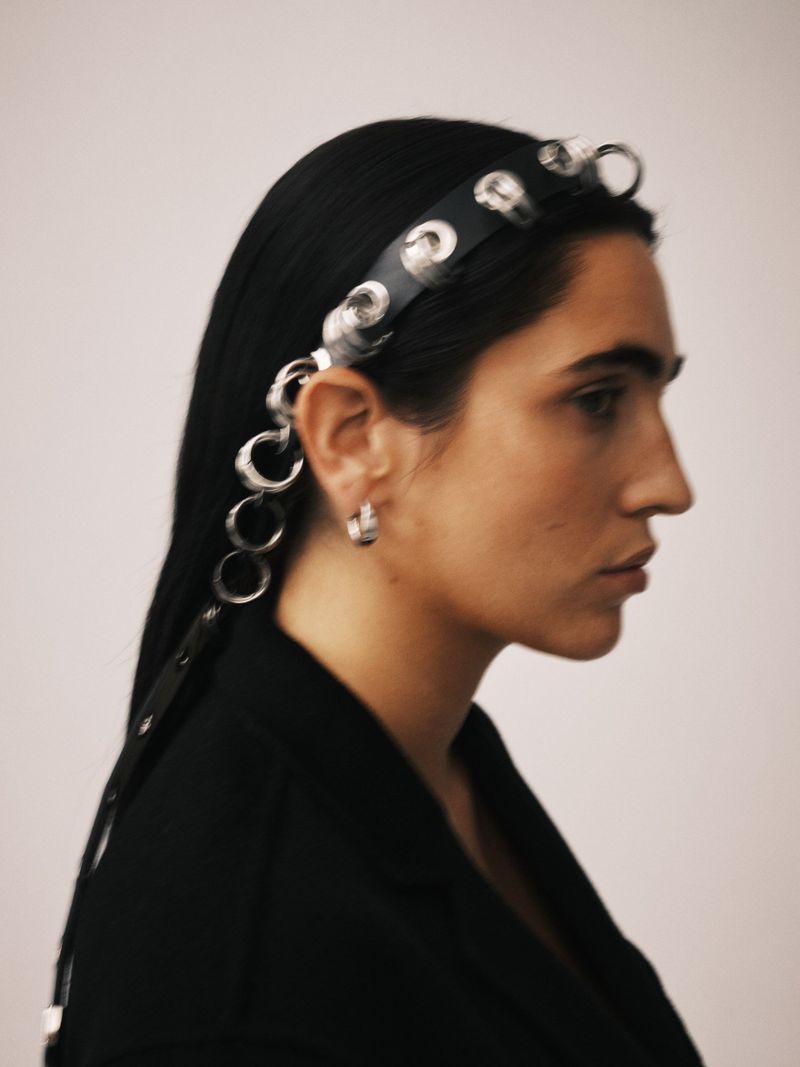 Profile shot of woman with long black hair wearing black blazer and eyelet headband against a light neutral studio backdrop.