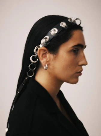 Profile shot of woman with long black hair wearing black blazer and eyelet headband against a light neutral studio backdrop.