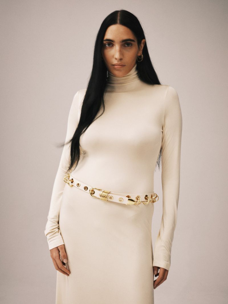 Model Valentine Valero in an off-white turtleneck dress and gold-hardware belt against a neutral studio backdrop.