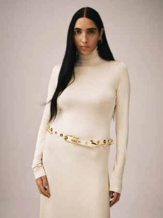 Model Valentine Valero in an off-white turtleneck dress and gold-hardware belt against a neutral studio backdrop.