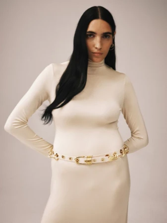 Adult female model poses in front of light background wearing a long sleeve, cream-colored dress, and a gold and cream belt.