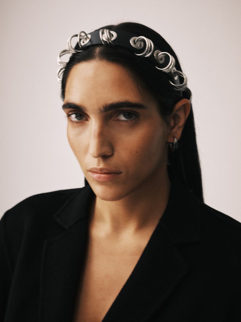 Close-up studio portrait of a dark-haired woman wearing a black jacket and designer headband with a serious expression against an off-white backdrop.