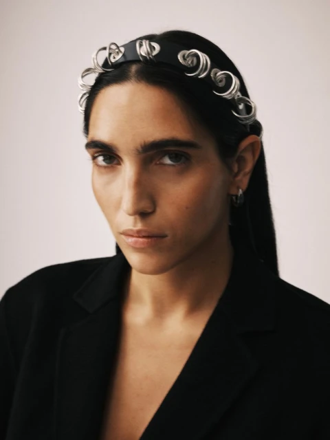 Close-up studio portrait of a dark-haired woman wearing a black jacket and designer headband with a serious expression against an off-white backdrop.