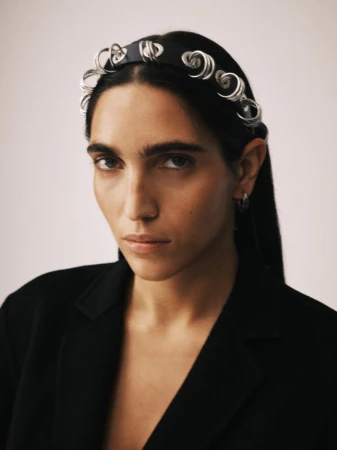 Close-up studio portrait of a dark-haired woman wearing a black jacket and designer headband with a serious expression against an off-white backdrop.
