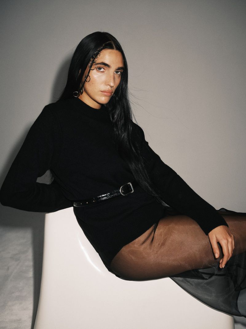 An adult woman poses in a photo studio wearing a black sweater with a belt and sheer black tights, seated on a white chair.