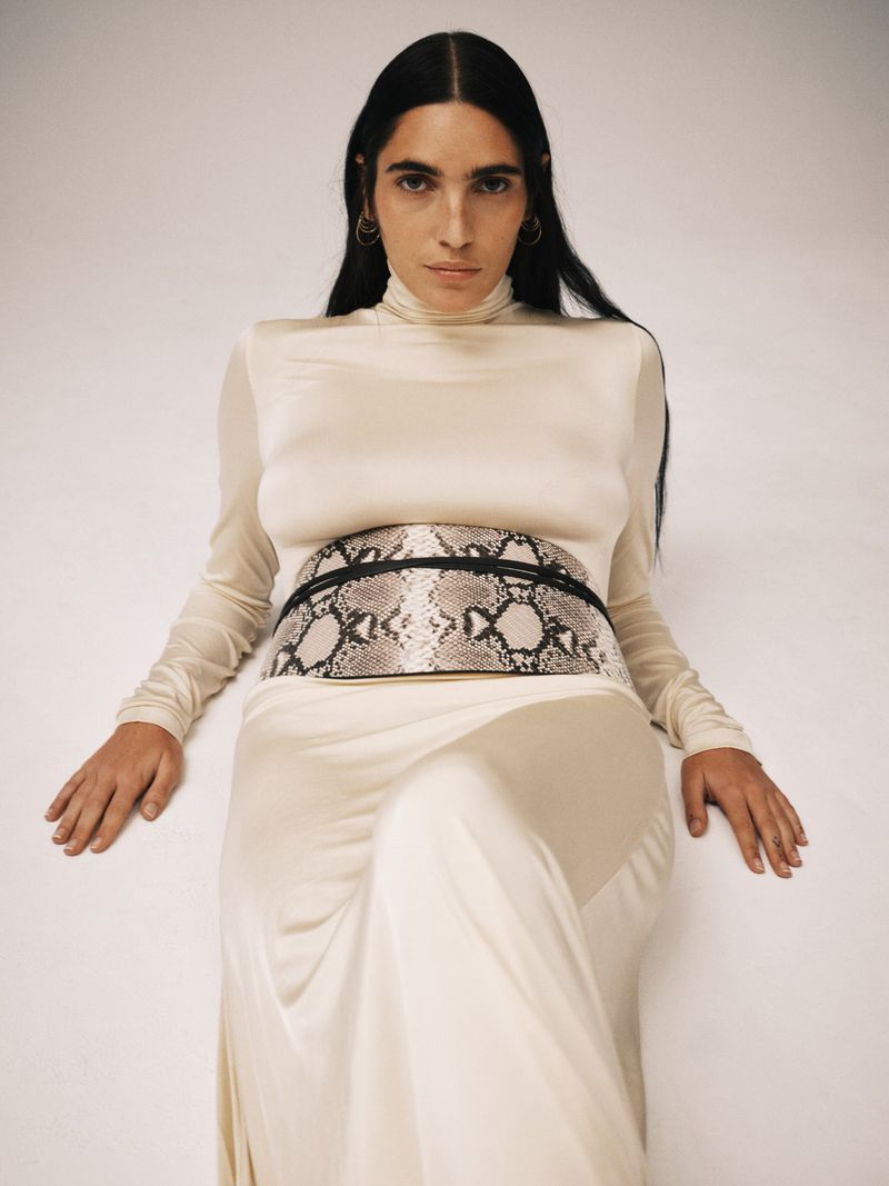 Woman in a cream-colored dress and snakeskin-print belt poses in front of a solid white backdrop in a fashion studio shoot.