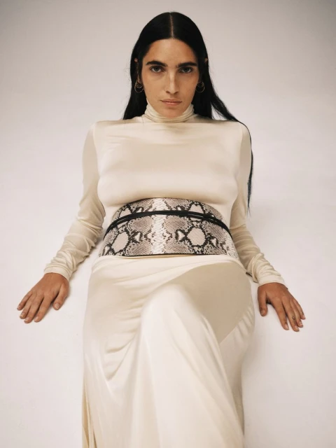 Woman in a cream-colored dress and snakeskin-print belt poses in front of a solid white backdrop in a fashion studio shoot.