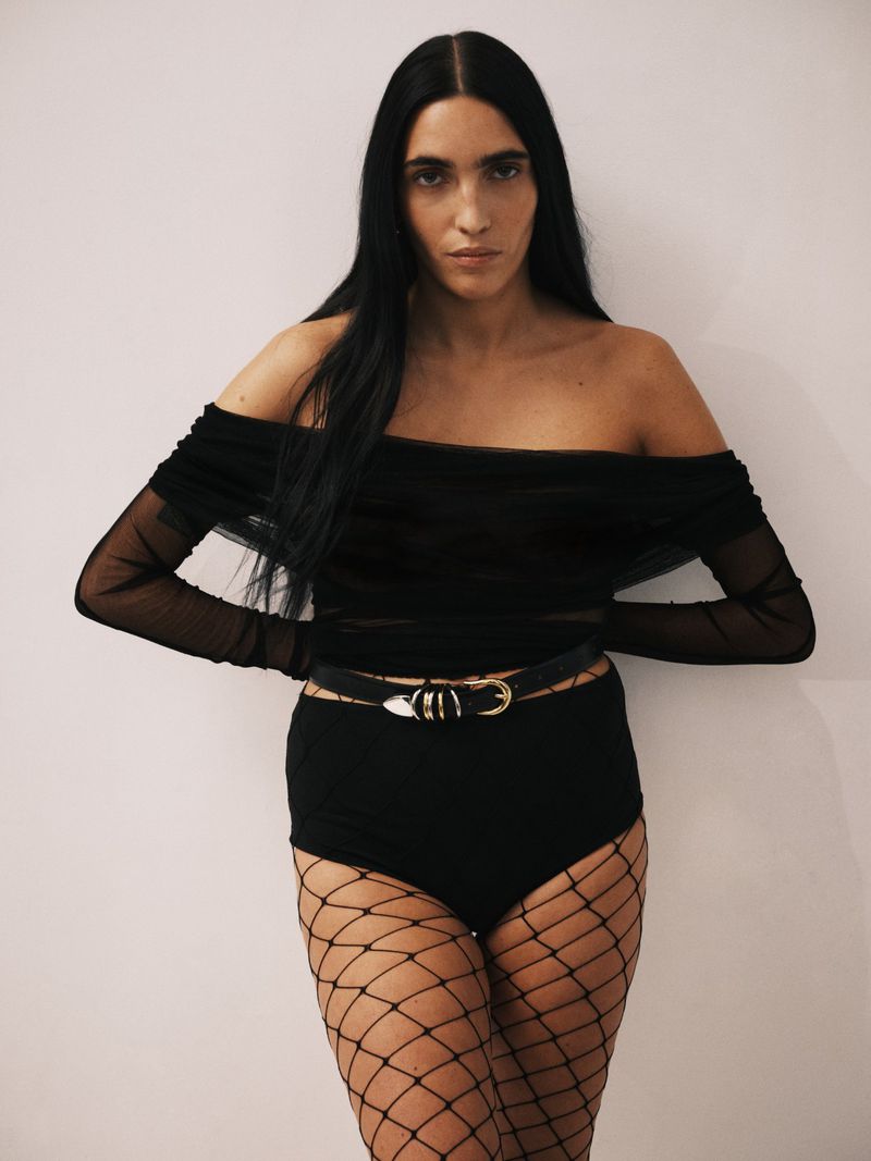 Woman in black mesh top and fishnet stockings posing against a neutral background. Full-length fashion portrait. Studio lit.