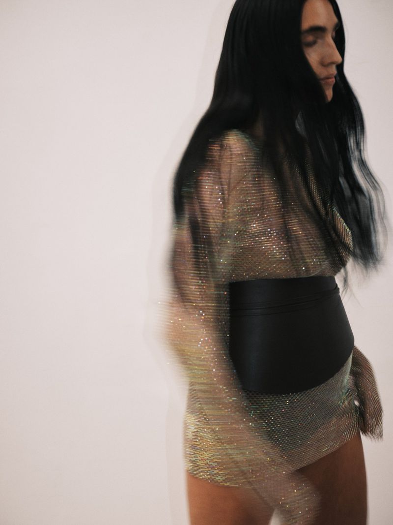 Fashionable woman with long dark hair wears a shimmering dress and black belt. Blurred motion in studio against a white backdrop.